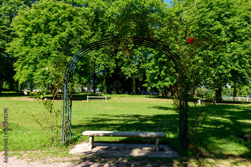 Fotografie A rustic stone bench sits beneath a wrought iron arch entwined with climbing roses, surrounded by lush green grass and tall leafy trees in the sunlight at Saint-Honore-les-Bains