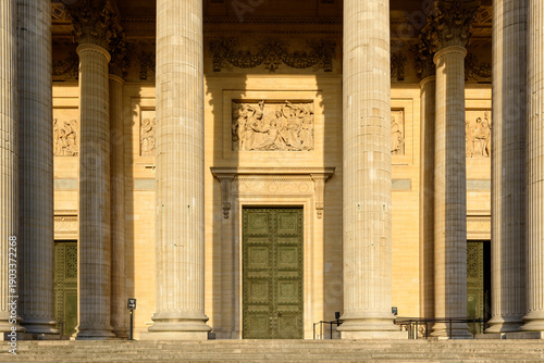 Tableau sur toile Close-up of the Pantheon's grand entrance in Paris, featuring tall neoclassical columns, ornate green doors, and sculpted stone reliefs illuminated by warm afternoon sunlight