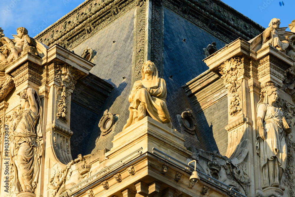 Fototapeta premium Golden evening light highlights a group of detailed stone sculptures and ornate reliefs at the corner rooftop of the Louvre palace in Paris, France. The renaissance architecture features intricate