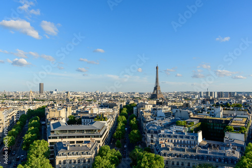 Expansive panoramic view over Paris featuring the Eiffel Tower rising above classic city rooftops and tree-lined avenues under a bright blue sky with scattered clouds.