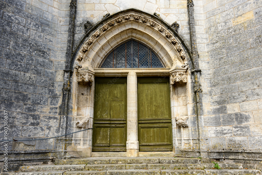 Fototapeta premium A weathered green double door set within a pointed stone archway forms the entrance to an old church in Pouilly sur Loire. Intricate Gothic details and aged masonry create a sense of heritage and