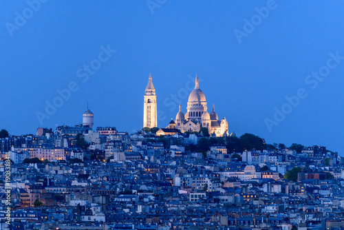 Bild auf Leinwand The illuminated domes and bell tower of Sacre Coeur Basilica rise above the densely packed rooftops of Montmartre at dusk, set against a vibrant blue evening sky