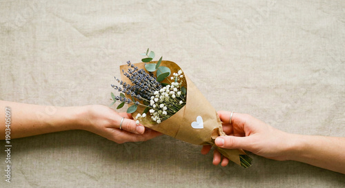 Close up view of two hands giving a rustic flower bouquet wrapped in brown paper with a small white heart sticker, lovely art of giving