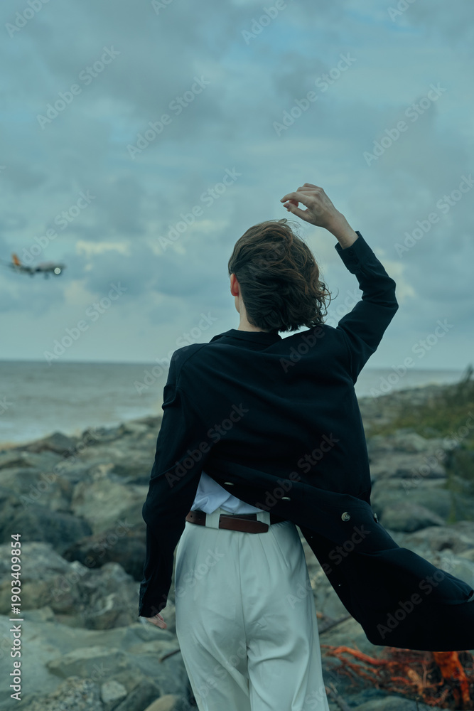 Fototapeta premium Coastline scene with figure in dark coat walking away on a rocky shore, wind blowing, white trousers, ocean backdrop and moody sky, suggesting adventure and contemplation.