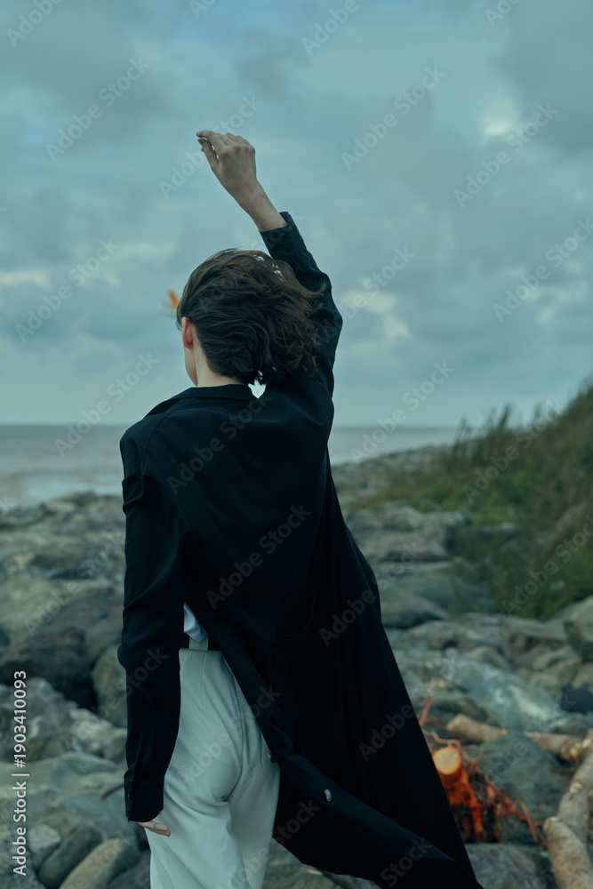 Fototapeta premium Woman stands on a rocky coast, wind in a dark jacket, raising a hand toward the sky as sea and clouds meet the horizon in a dramatic outdoor scene.