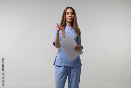 Fotografie Female caucasian young nurse in light blue scrubs holding papers and pointing