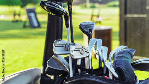 Billede på lærred Golf clubs are ready in a bag beside a green field where people play golf on a s