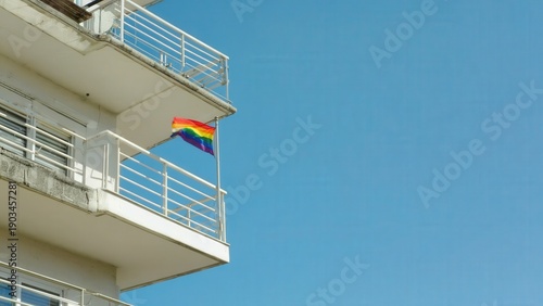 A rainbow flag waves from a balcony of a modern building against a clear blue sky. The scene symbolizes pride and inclusivity.