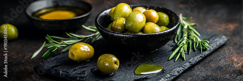 Fresh green olives in a black bowl with extra virgin olive oil and rosemary sprigs on a dark rustic stone background. Perfect for Mediterranean diet and gourmet cooking.