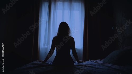 black silhouette upset unhappy woman sitting on bed dark room, mood depression sadness. Sad lonely Girl feels bad after party migraine suffers from headache. window curtains white fabric fly wind blow