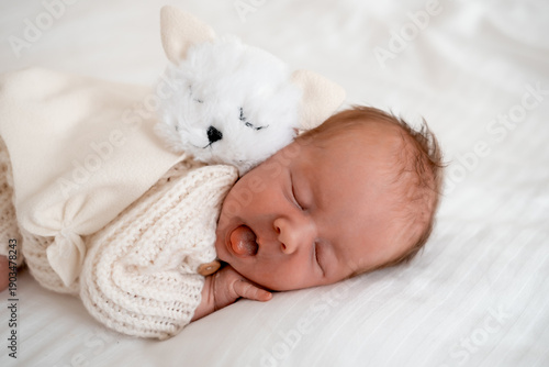 newborn baby sleeping on a white bed at home with a soft toy, close-up portrait of a newborn baby sleeping in a white knitted bodysuit, space and room for text