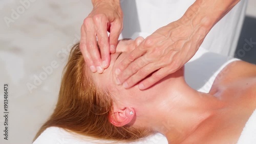 A redheaded woman lies on a white towel as veined hands apply oil and gentle facial and scalp massage, with a diamond stud earring, in bright outdoor light.