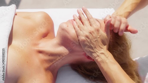 Overhead close up shows rhythmic hand motions on jawline, cheeks, and temples, bright daylight, white towel, floral stud earring, high key look, shallow depth.