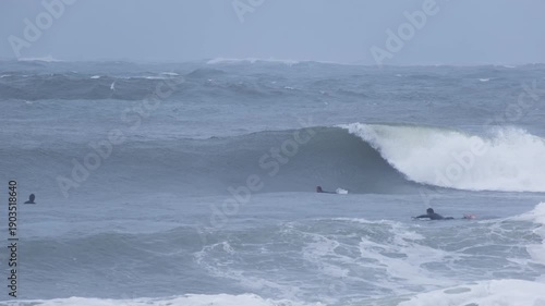 Surfers in wetsuits paddle and ride powerful breaking waves in rough sea conditions. Outdoor adventure and extreme water sport scene.
