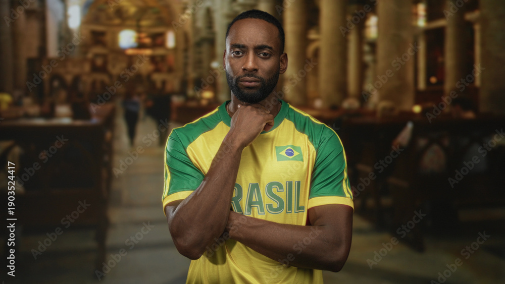 Fototapeta premium Man with hand on neck, wearing brazil soccer jersey and folded arms, standing in a church building amid pews and pillars; pain reflection.