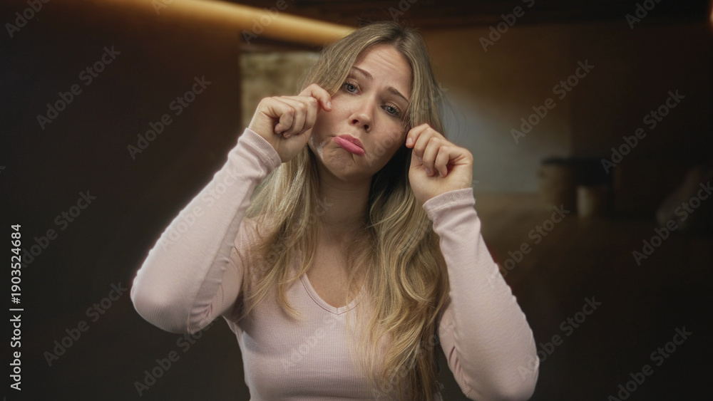 Fototapeta premium Blonde woman wearing pink top wipes tears at her eyes beside wooden table under soft warm light in room; sadness sorrow vulnerability.