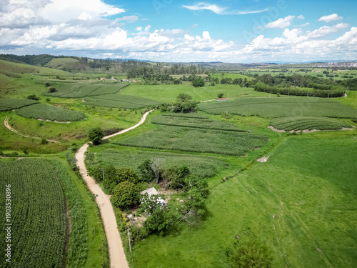 Konstfotografi Vista aérea com drone de uma plantação de milho cercada por morros verdes e área rural, formando uma paisagem agrícola ampla e cinematográfica sob céu nublado