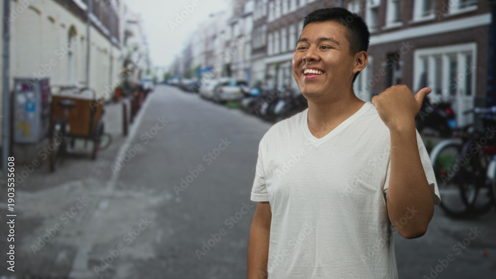 Fototapeta premium Hispanic young man pointing thumb over shoulder on street while smiling; casual confidence happiness.