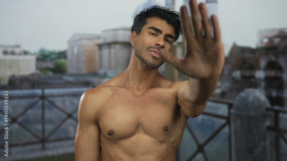 Obraz premium Young hispanic shirtless man raising palm as stop sign before roman ruins building; refusal defiance determination.