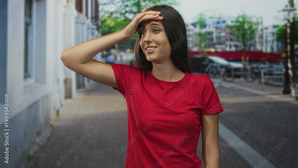 Fototapeta premium Young hispanic woman in red shirt with hand on forehead on a city street, palm to brow gesture; embarrassment.