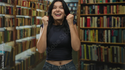 Foto Woman pumping fists among bookshelves in library while wearing navy top and jeans, smiling broadly; excitement