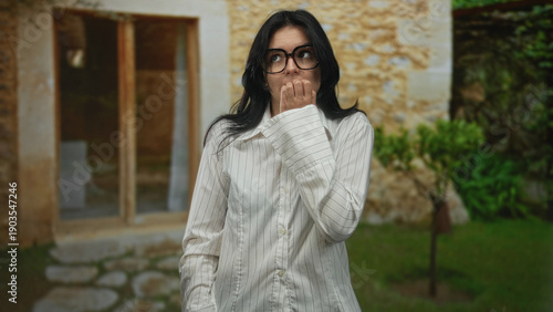 Tableau sur toile Woman biting nails gesture with hand to mouth in front of rustic stone building outdoors, wearing glasses and striped shirt; uncertainty
