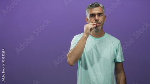 Canvas Print Man with grey hair in mint green tshirt pinching fingers to lips in studio, gesture suggests silence or a small pinch near mouth; quiet contemplation