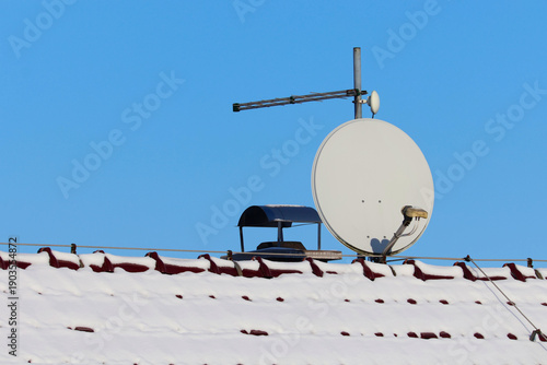 Satellite dish and TV antenna on a snowy house roof against clear blue sky, winter technology, telecommunication in cold weather, home equipment.