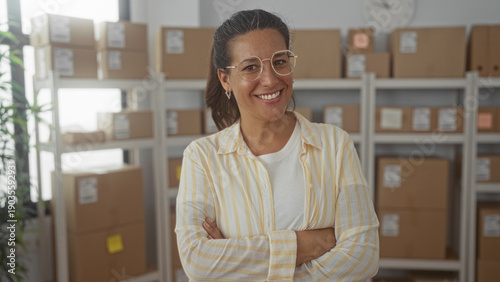 Foto Woman with crossed arms among cardboard boxes in a storage building, wearing glasses and neutral expression; quiet determination