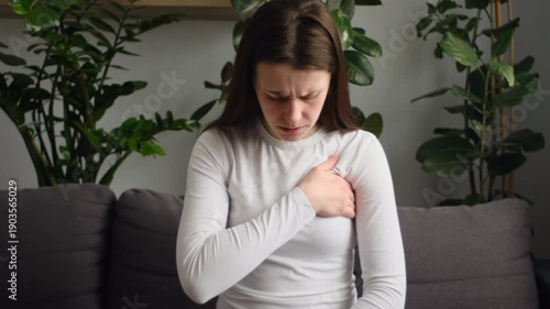 Unhealthy sad young caucasian woman touching her breast sitting alone on couch at home. Breast cancer self check. Healthcare and medical concept