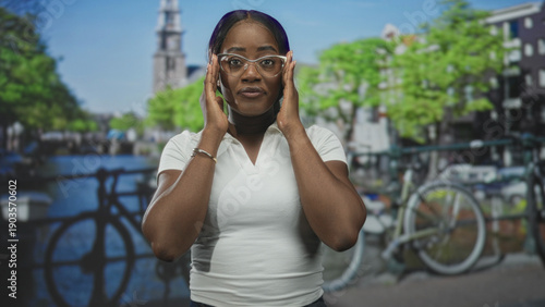 Photography Woman wearing glasses and white polo gives double thumbs up and briefly adjusts glasses in a studio with canal bridge backdrop; confidence