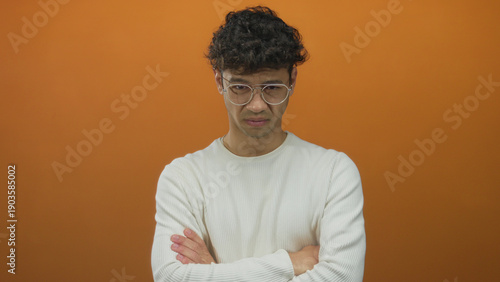 Man frowning with bespectacled face and arms crossed under chin in studio wearing white sweater; disgust.