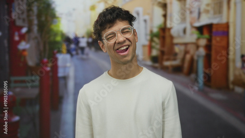 Foto Man wearing glasses with tongue out winking in colorful urban street scene with blurred pedestrians; playful mood