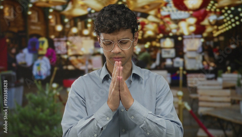 Εκτύπωση καμβά Man wearing glasses with clasped hands in prayer on street market under hanging lights; serenity
