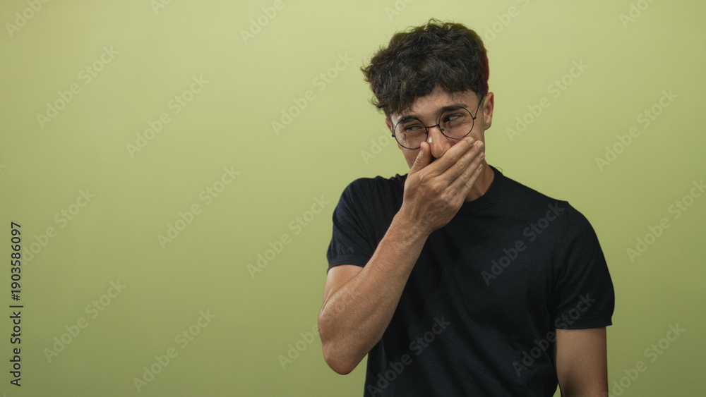 © Krakenimages.com - Young hispanic man wearing glasses and black t shirt covers mouth with hand in olive studio; amusement shyness. © Krakenimages.com - Young hispanic man wearing glasses and black t shirt covers mouth with hand in olive studio; amusement shyness.