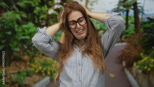 Foto Woman with glasses covers ears and holds head amid lush green forest pathway under sunlight; stress