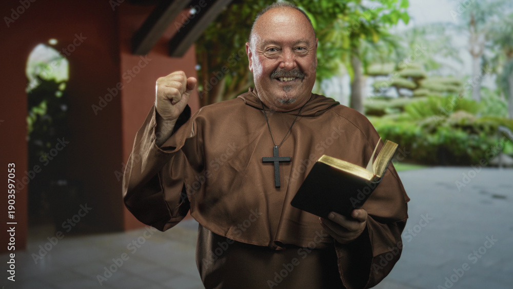 Fototapeta premium Man monk in brown robe holds open bible, raises right hand and smiles in a building courtyard; joy blessing.