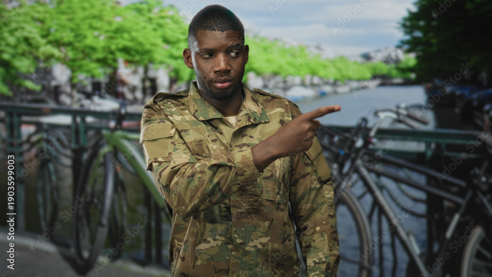 Fototapeta premium Man soldier in camouflage uniform with finger to lips for silence beside parked bicycles on a street canal bridge; secrecy duty.