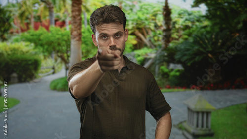 Man points finger at camera in park path with palm trees and a stone lantern beside a paved walkway, visible hand and forearm gesture; confidence challenge persuasion.
