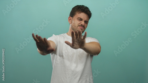 Young hispanic man palms out stop gesture, bare hands and face visible, studio teal backdrop; repulsion refusal avoidance.