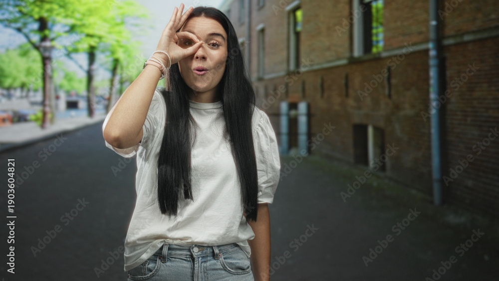 Fototapeta premium Young hispanic woman standing on a city street by a brick building, making ok sign with hand over eye while smiling in a white t shirt; playful curiosity.