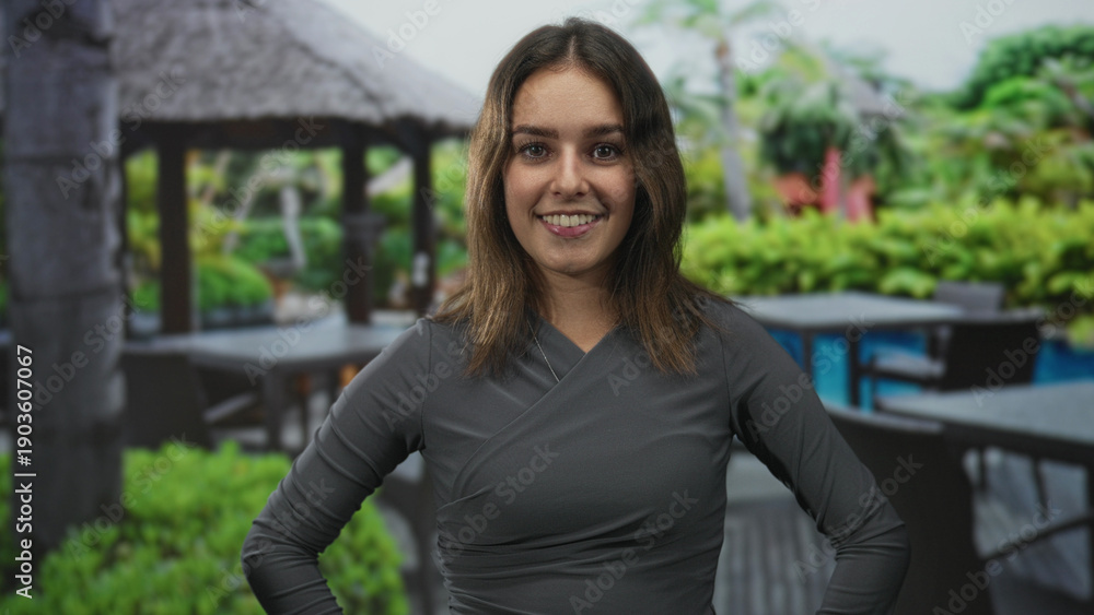 Fototapeta premium Woman smiling with hands on hips and visible face at resort poolside gazebo and outdoor patio; confidence optimism.
