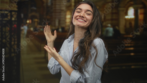 Konstfotografi Young hispanic woman gestures with both hands while smiling inside a historic church building under soft daylight; serenity