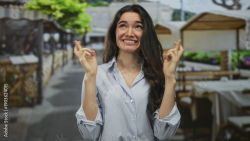 Woman crossing fingers at a sunlit restaurant terrace with wooden chairs and potted plants lining a stone walkway; hopeful anticipation.