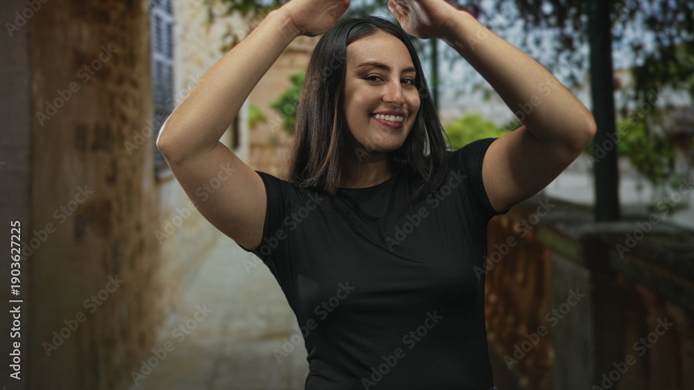 Fototapeta premium Woman smiling with hands above head revealing bare arms on a narrow street near stone buildings; joy casual movement.