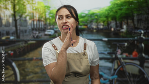 Photography Woman in apron cups her hand to mouth while holding a pen on a street in amsterdam near a canal bridge; thoughtful focus