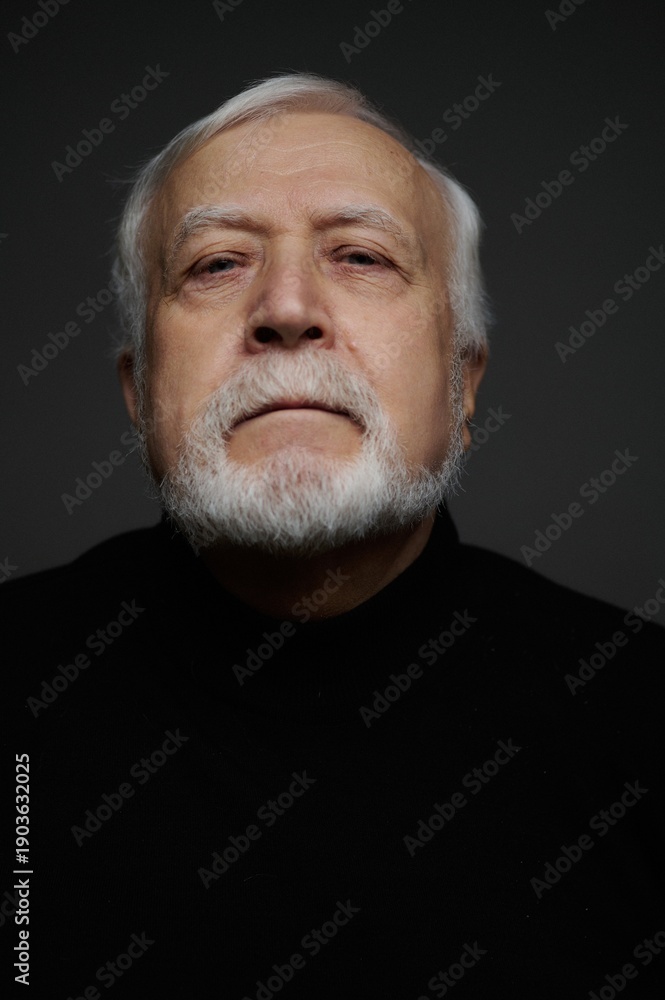Obraz premium Elderly man with white beard looks directly at the camera in a simple light setting against a dark background