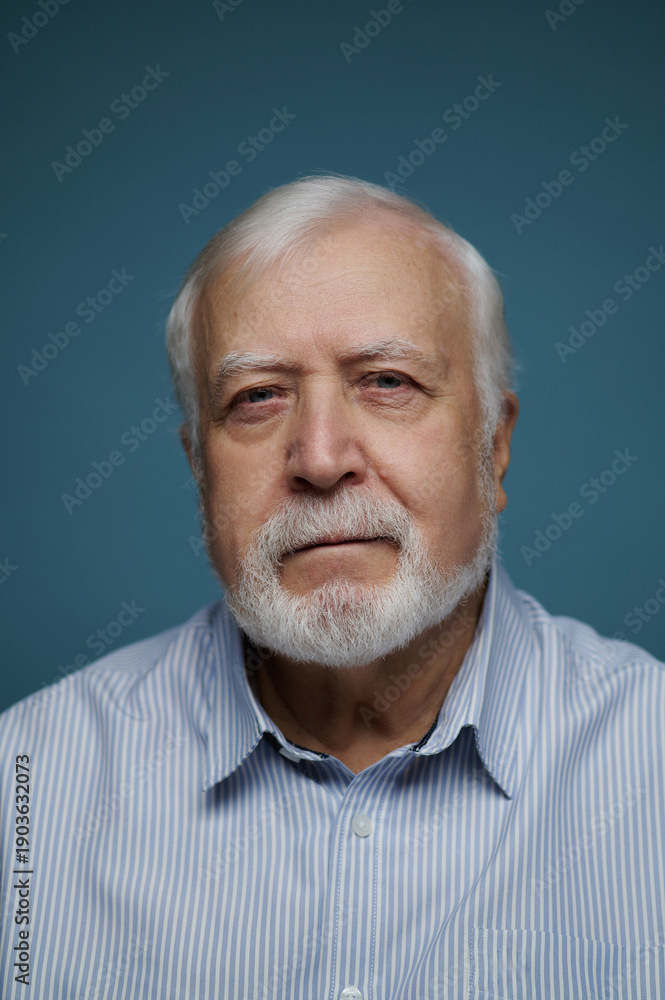 Fototapeta premium Elderly man with white beard wearing a blue striped shirt looking into the camera in a simple indoor setting