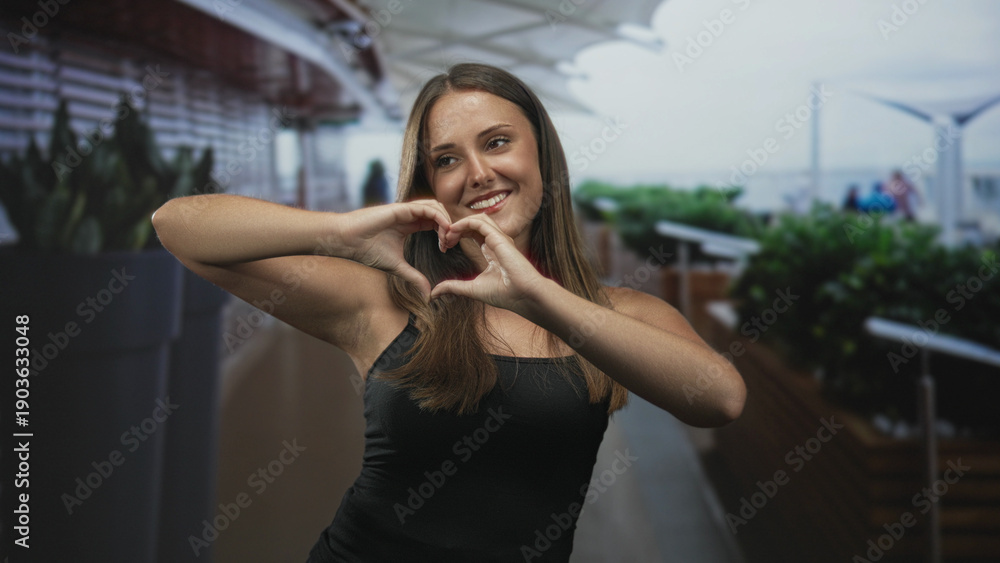 Fototapeta premium Young woman in black tank top forms heart with hands, smiling near large planters and railing on an airport terrace under canopy; warm affection.