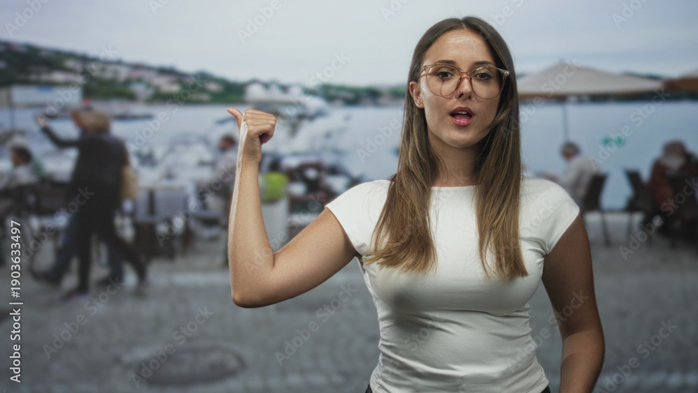 Fototapeta premium Young woman with glasses and white t shirt thumb pointing to marina and outdoor cafe seating at street waterfront; contentment.